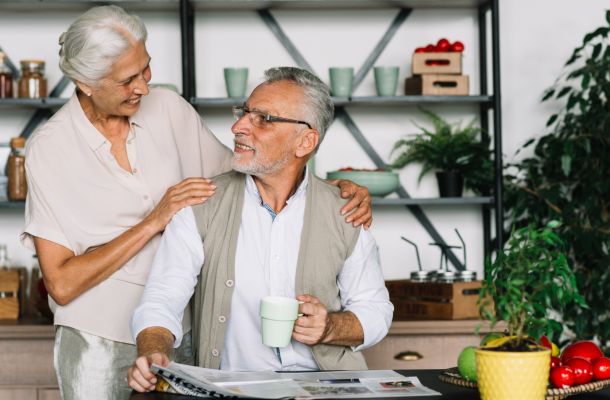 Smiling elderly couple in a kitchen, one holding a mug and newspaper, surrounded by plants and fresh produce.