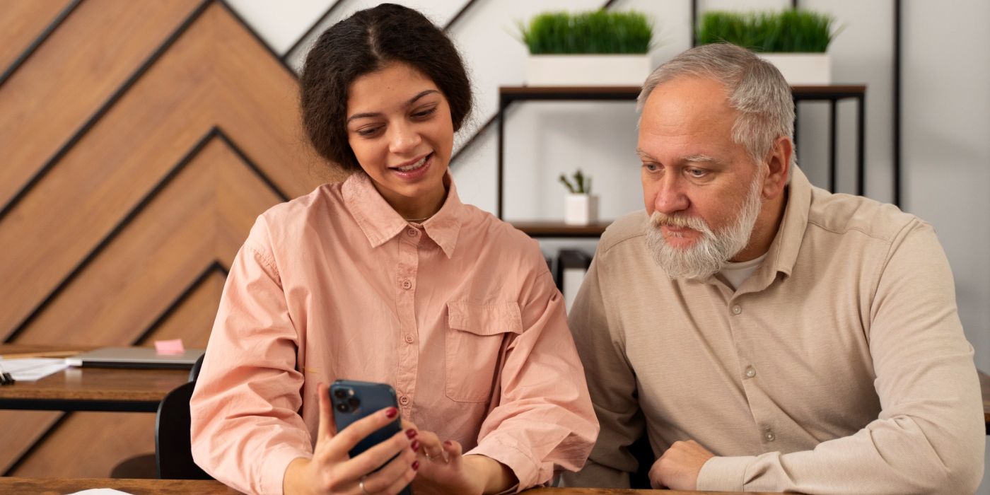 A young woman shows something on her phone to an older man as they sit together at a desk in a modern office.