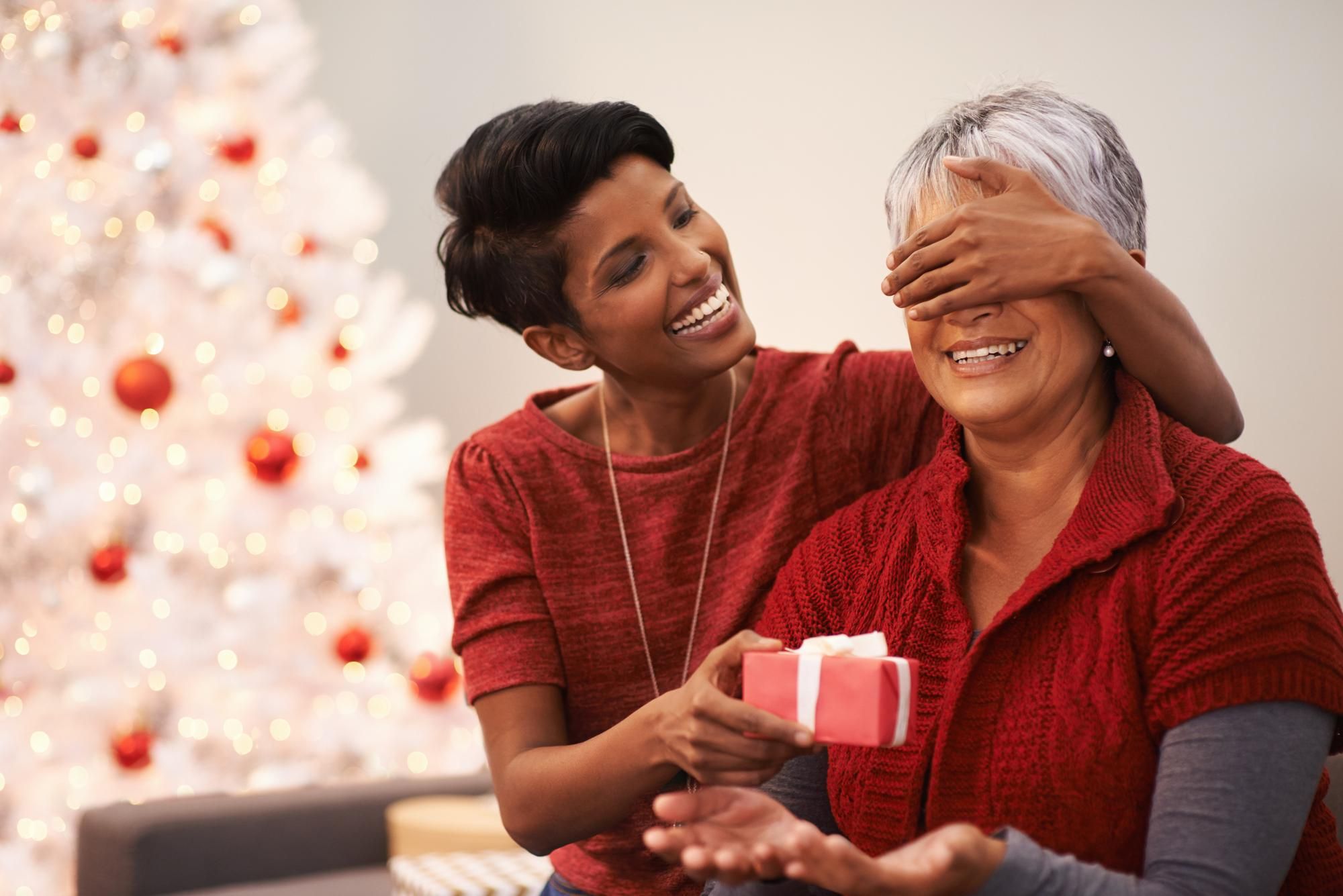 Young woman surprises smiling older woman with a gift in front of a decorated Christmas tree.