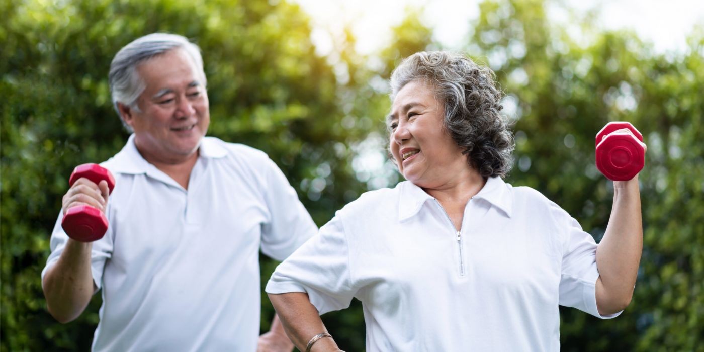 Smiling seniors in white shirts exercise outdoors with red dumbbells, enjoying a bright, green background.