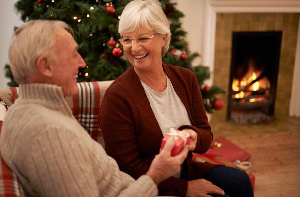 Smiling older couple exchanging a gift by a Christmas tree and fireplace.