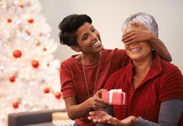 Young woman surprises smiling older woman with a gift in front of a decorated Christmas tree.