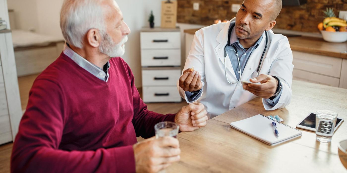 A doctor explains medication to an older man at a kitchen table; both hold glasses of water.