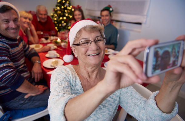 Smiling elderly woman in Santa hat taking a selfie with family at a festive table near a Christmas tree.