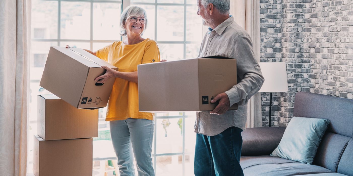 Smiling older couple holding moving boxes in a living room with stacked boxes and a large window.