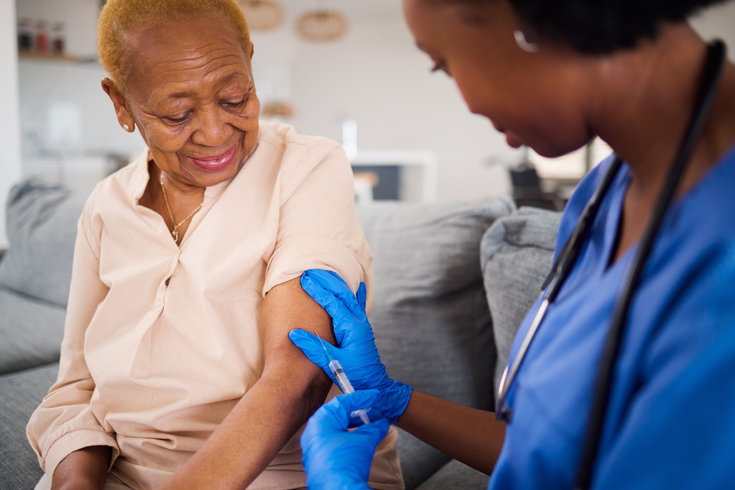 Smiling elderly woman receives a vaccine shot in her arm from a nurse wearing blue gloves.