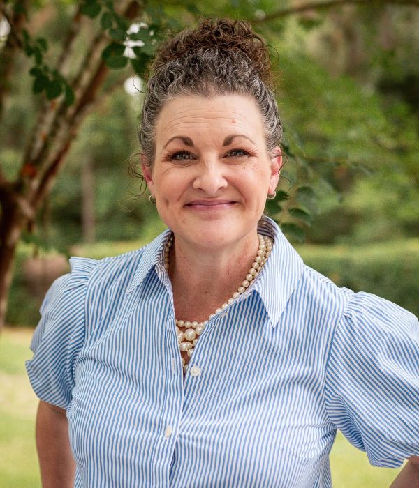 Smiling woman with curly hair in a striped shirt and pearl necklace standing outdoors with greenery.