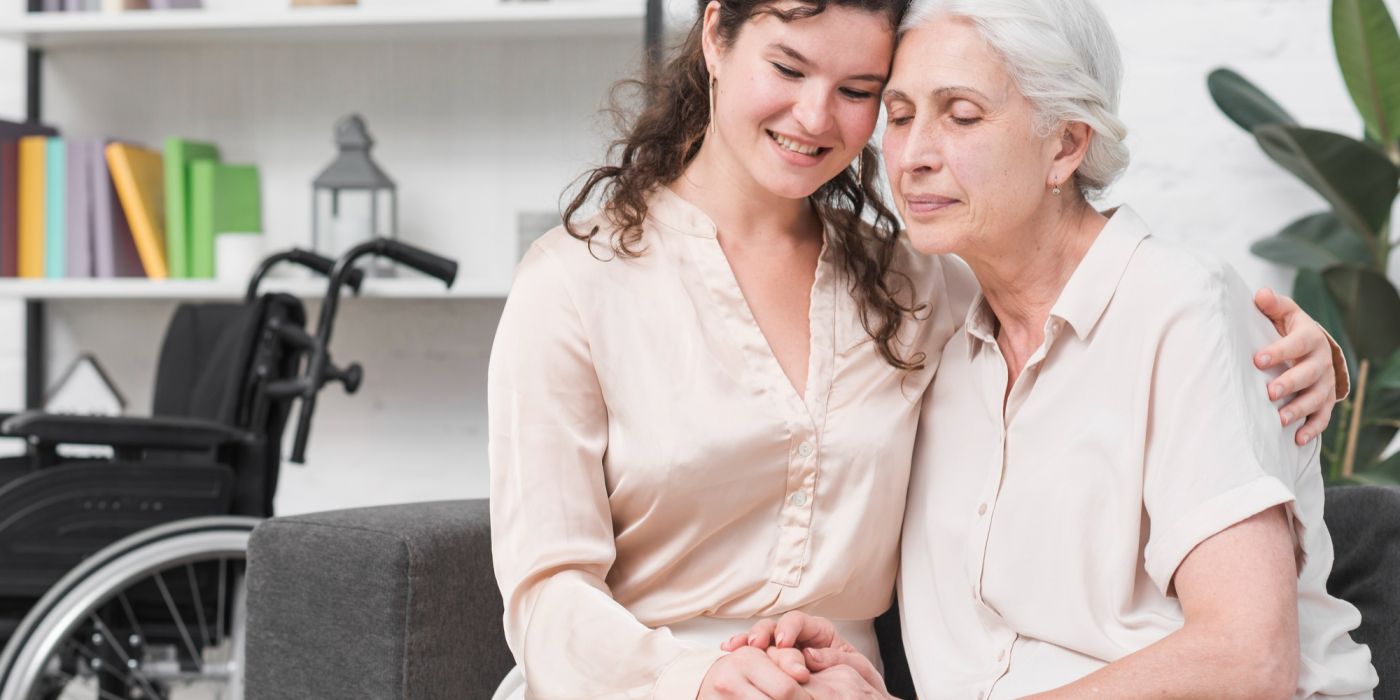 Young woman hugging elderly woman on couch, wheelchair in background, both smiling and holding hands.