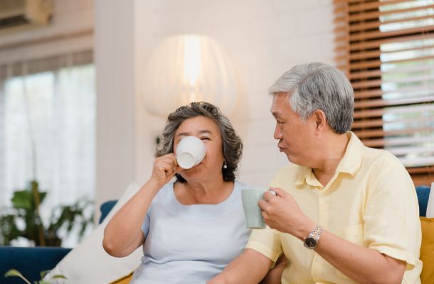 Older couple sitting on a couch at home, enjoying hot drinks and smiling together.