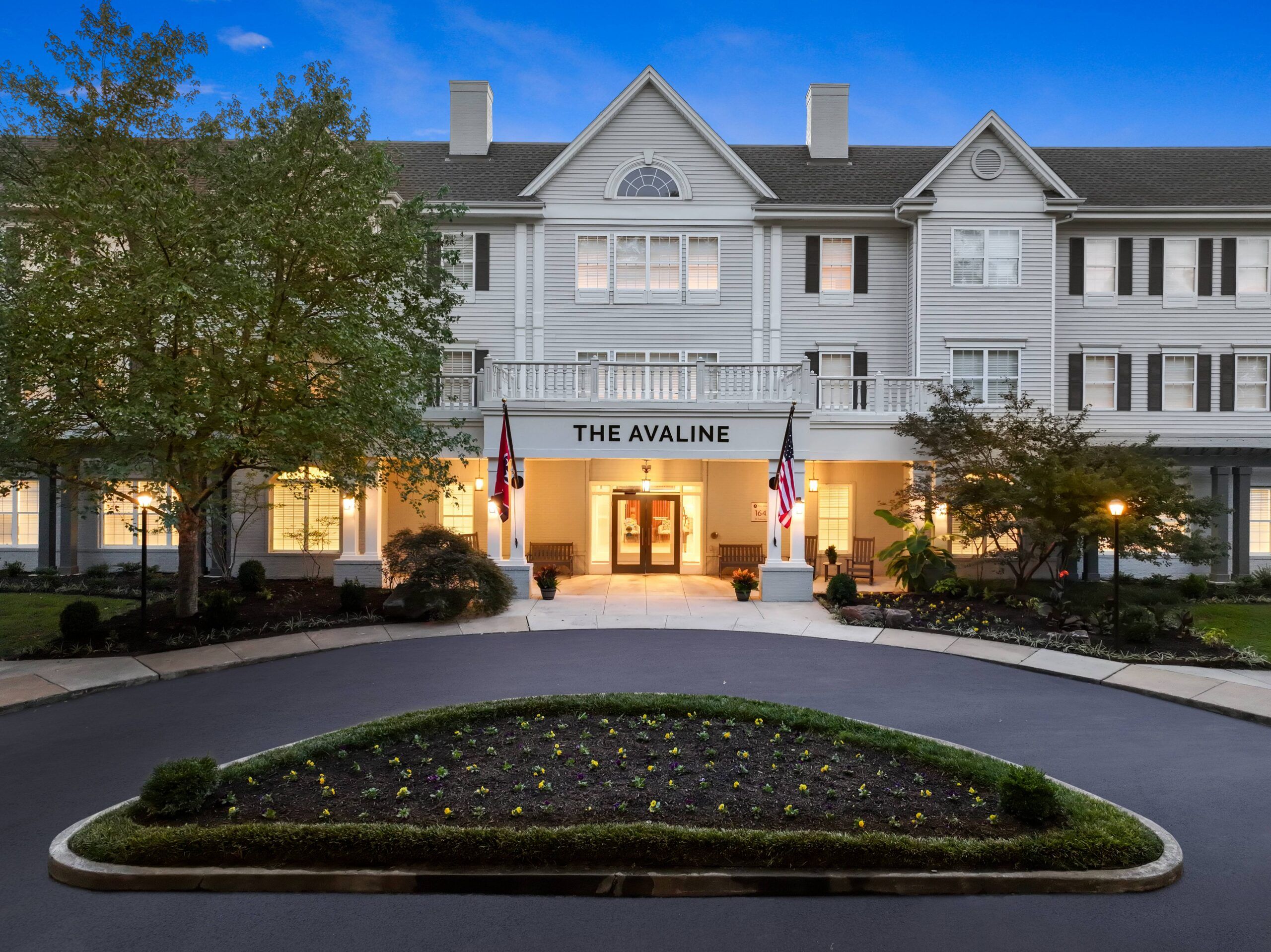 A large white senior living community named The Avaline with lit windows, two flags, and a landscaped driveway at dusk.