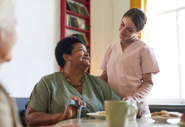 Smiling caregiver stands beside a seated woman, both sharing a warm interaction in a cozy room.