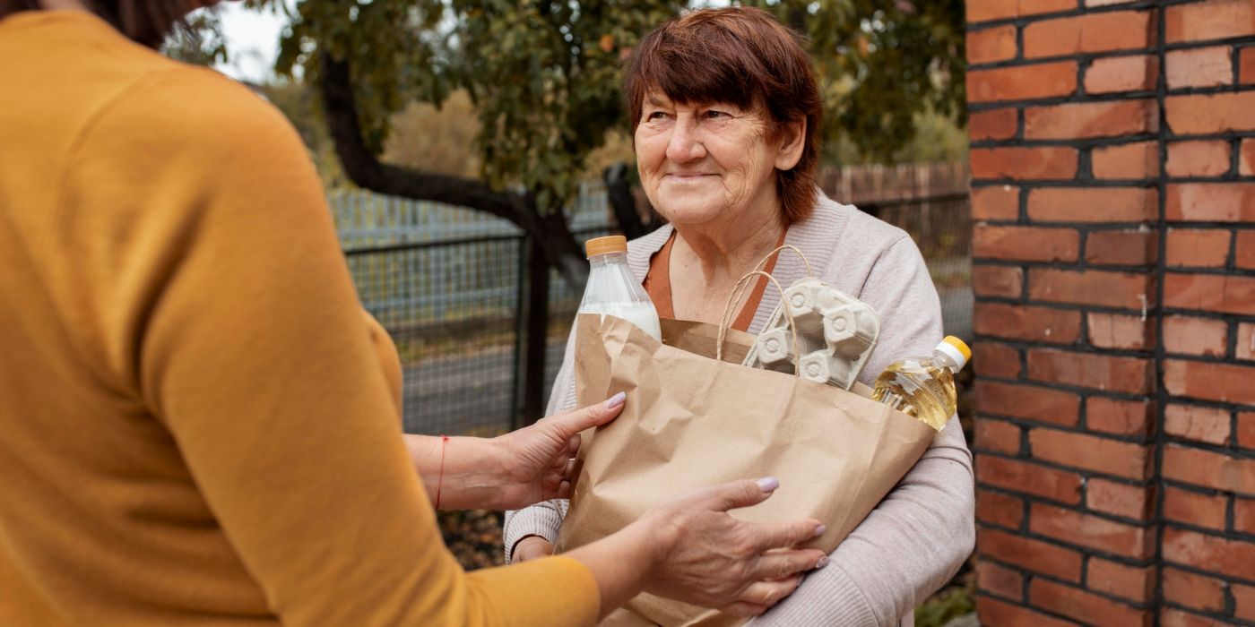 Elderly woman receiving a paper grocery bag from another person outdoors near a brick wall.