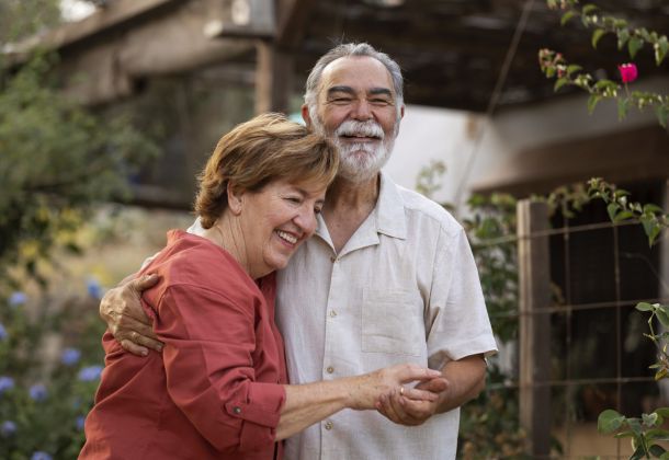 Smiling elderly couple hugging outdoors in a garden, both looking happy and relaxed.