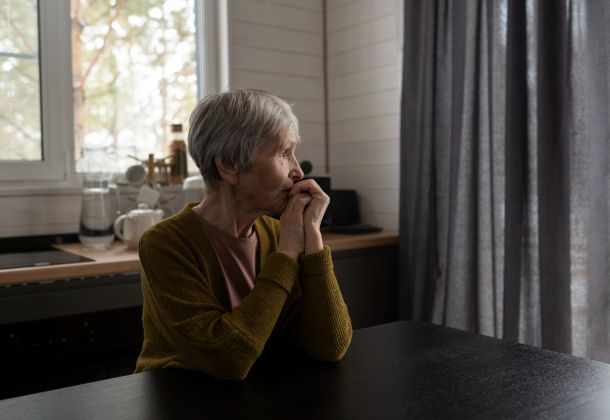 An elderly woman sits at a table, looking thoughtfully out a window in a softly lit kitchen.