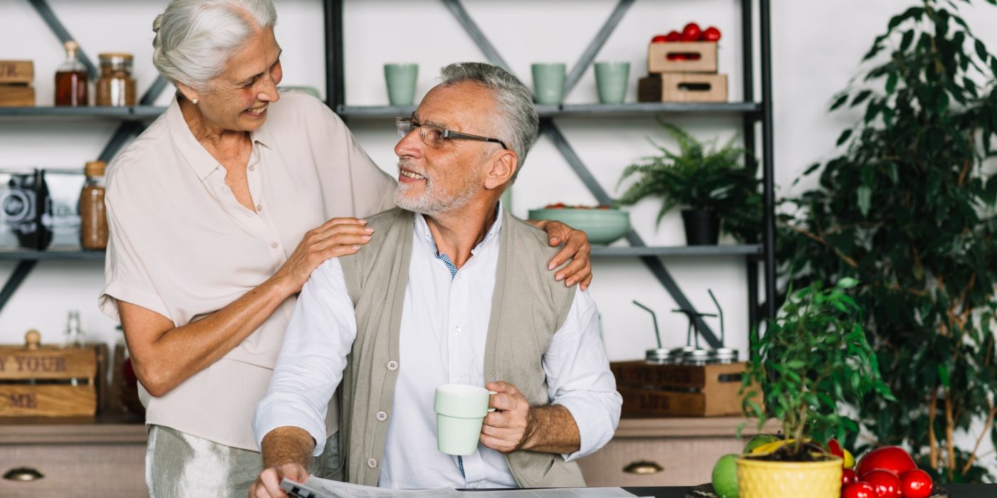 Smiling elderly couple in a kitchen, one holding a mug and newspaper, surrounded by plants and fresh produce.