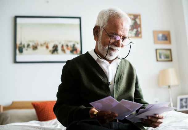 An elderly man with a white beard and glasses smiles while looking at old photographs in his bedroom.