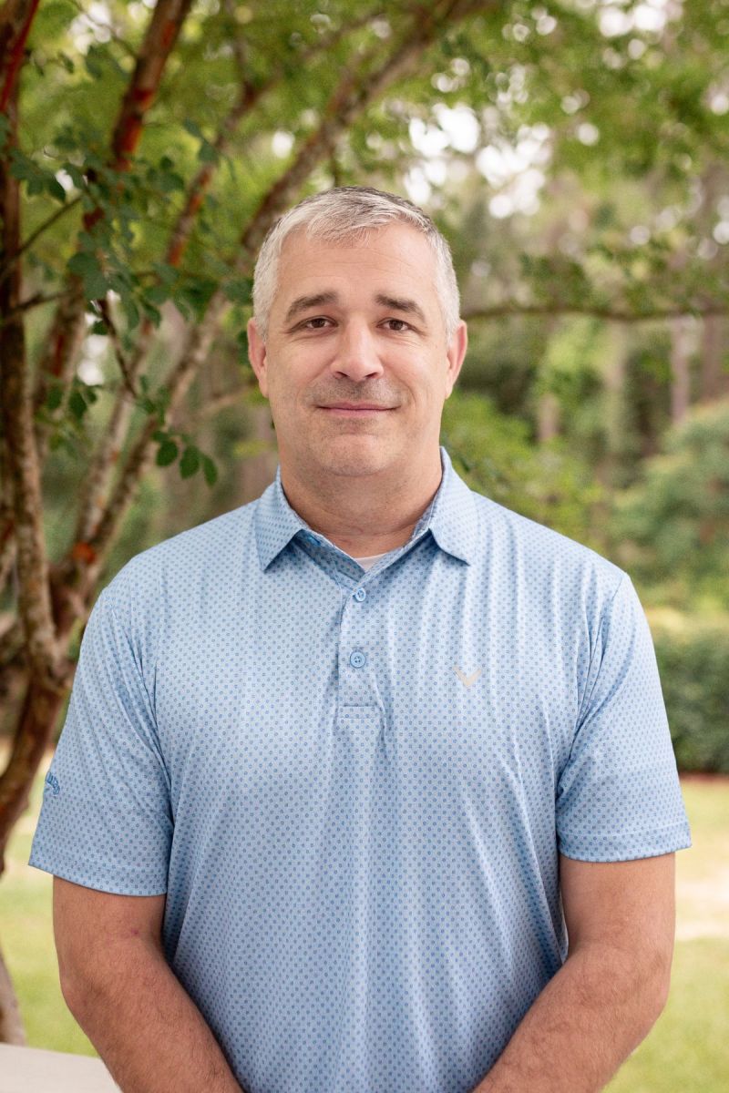 A man in a light blue polo shirt stands outdoors in front of green trees, smiling slightly at the camera.