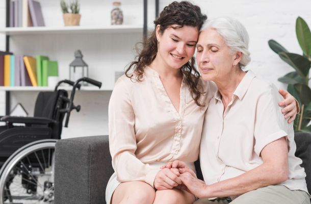 Young woman hugging elderly woman on couch, wheelchair in background, both smiling and holding hands.