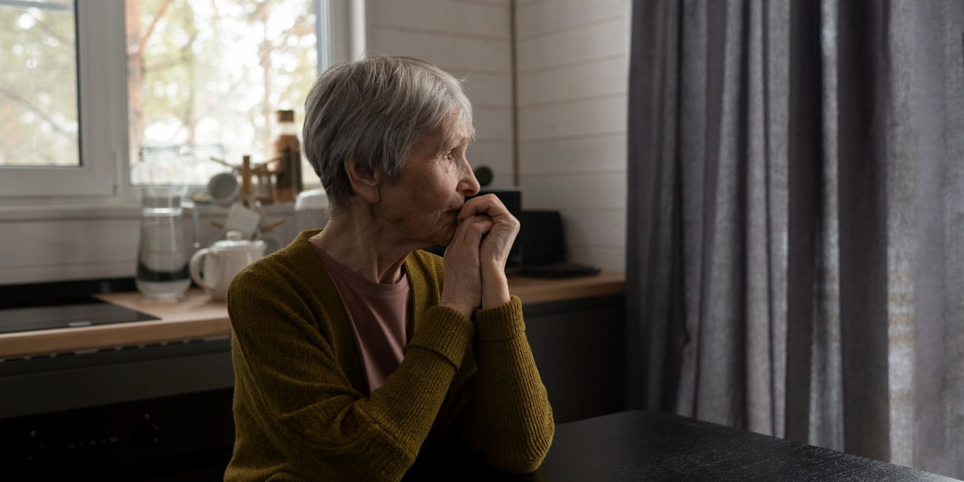 An elderly woman sits at a table, looking thoughtfully out a window in a softly lit kitchen.