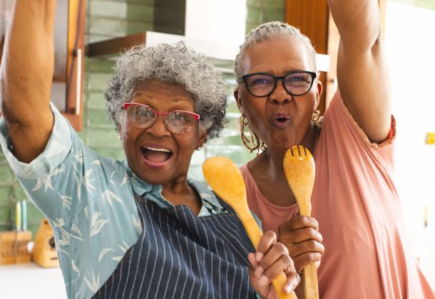 Two joyful older women in a kitchen, holding wooden utensils and smiling energetically at the camera.
