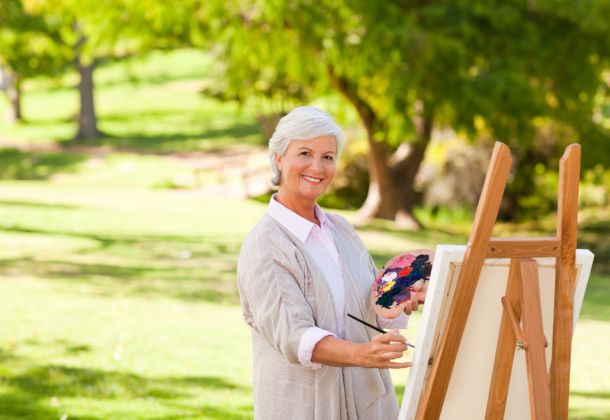 Smiling older woman painting at an easel outdoors in a park, holding a palette with colorful paint.