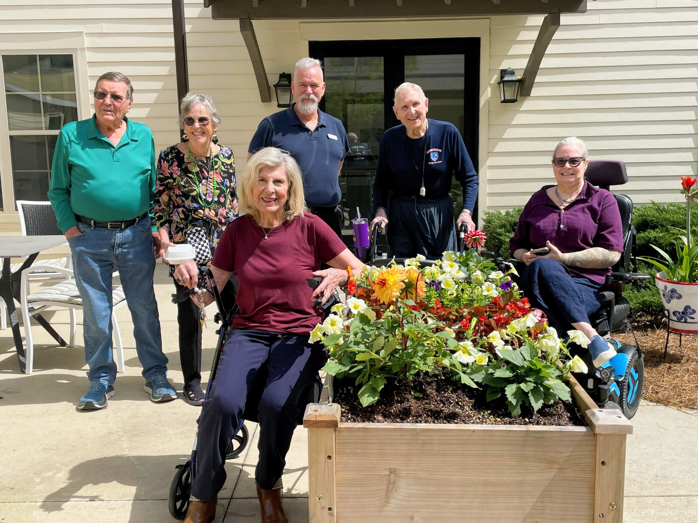 Six smiling seniors pose outdoors around a raised flower bed filled with colorful blooming flowers.