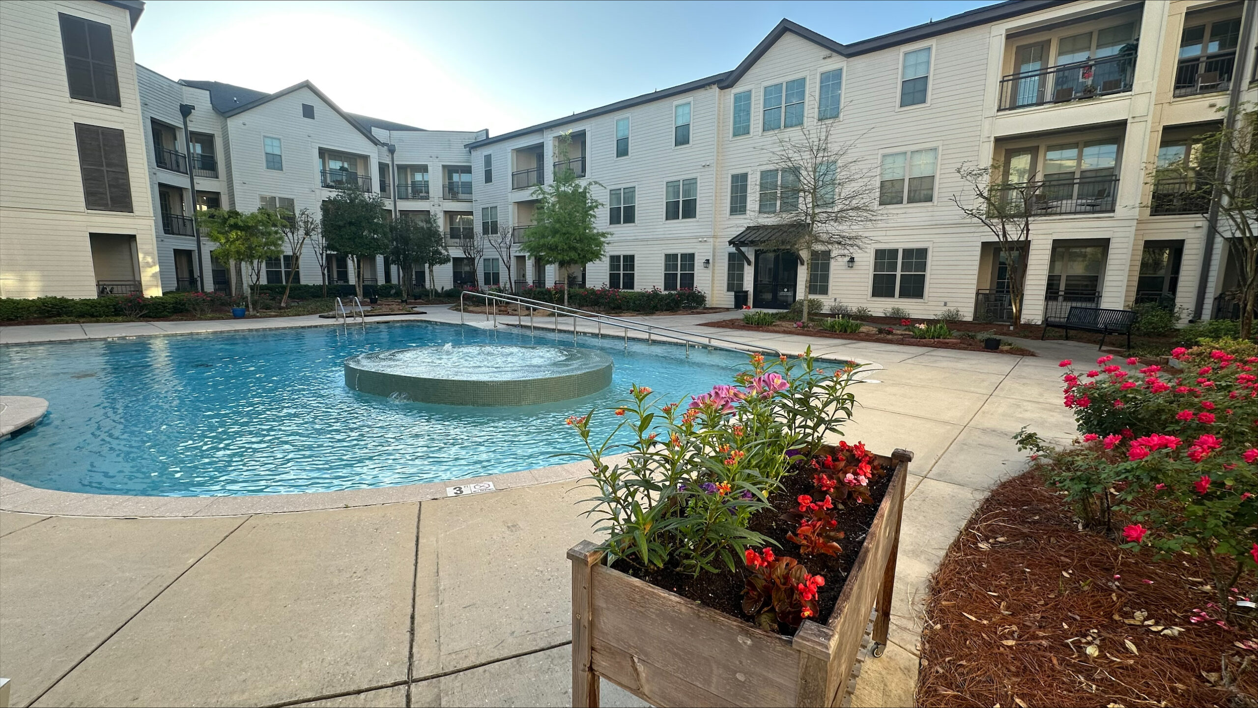 Outdoor swimming pool surrounded by apartments, flowers, and landscaping on a sunny day.